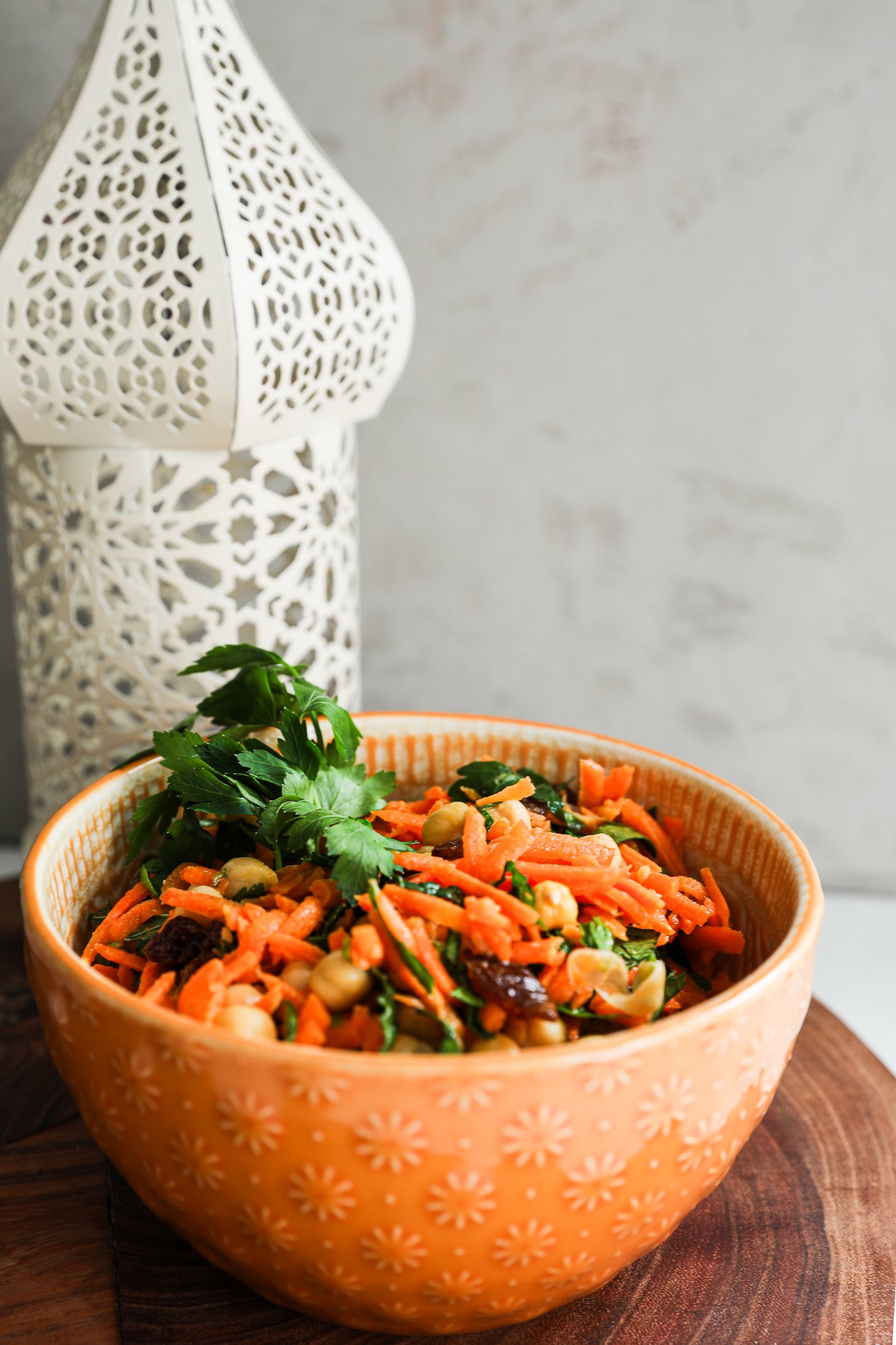 Perspective image of an orange bowl of grated carrot salad with raisins, chickpeas and herbs.