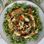 A large plate of a bed of chopped kale, topped with roasted charred sweet potato wedges doused in a creamy dressing and covered with chopped pistachios and fresh dill.