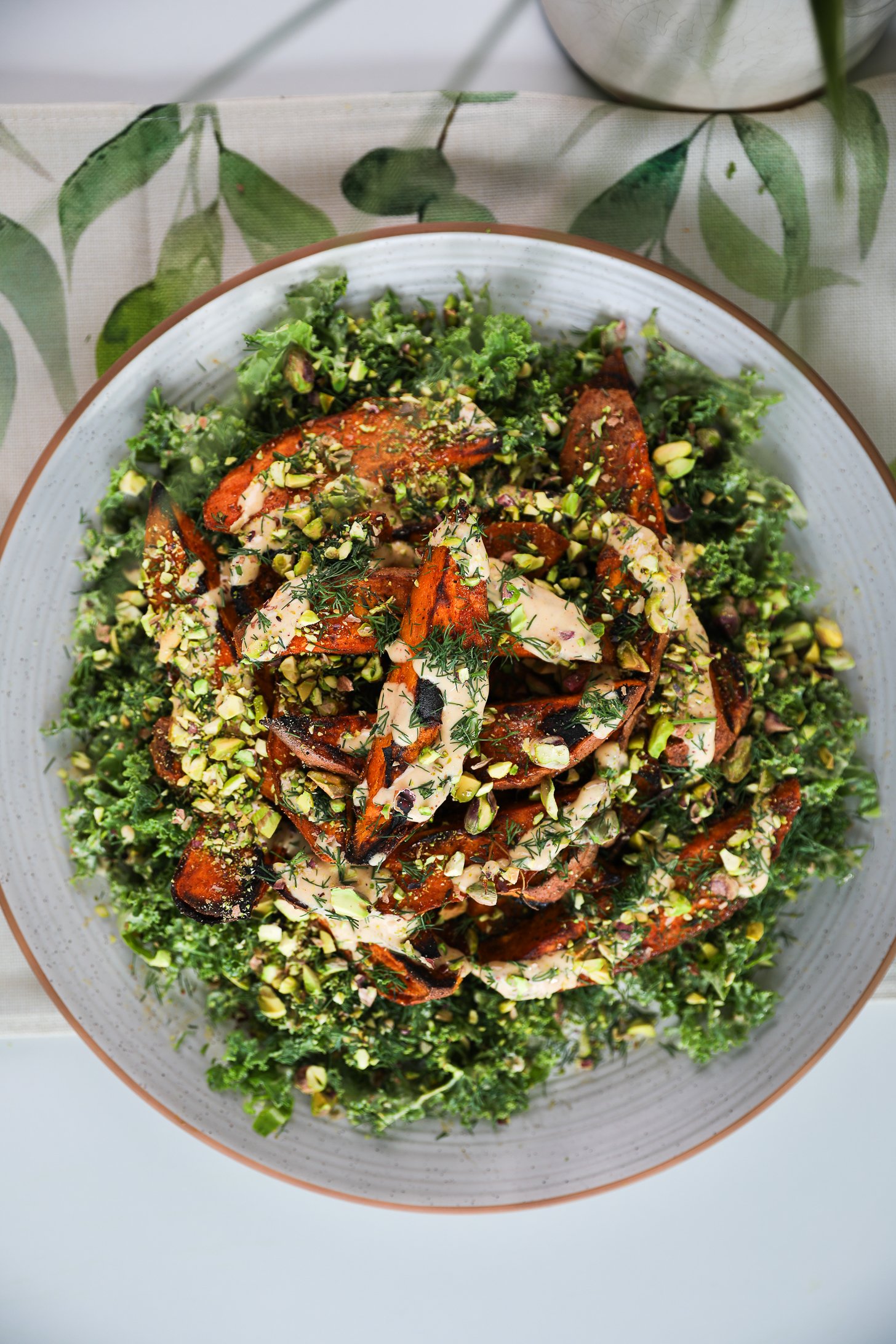A large plate of a bed of chopped kale, topped with roasted charred sweet potato wedges doused in a creamy dressing and covered with chopped pistachios and fresh dill.