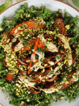 A close up image of a large plate of a bed of chopped kale, topped with roasted charred sweet potato wedges doused in a creamy dressing and covered with chopped pistachios and fresh dill.