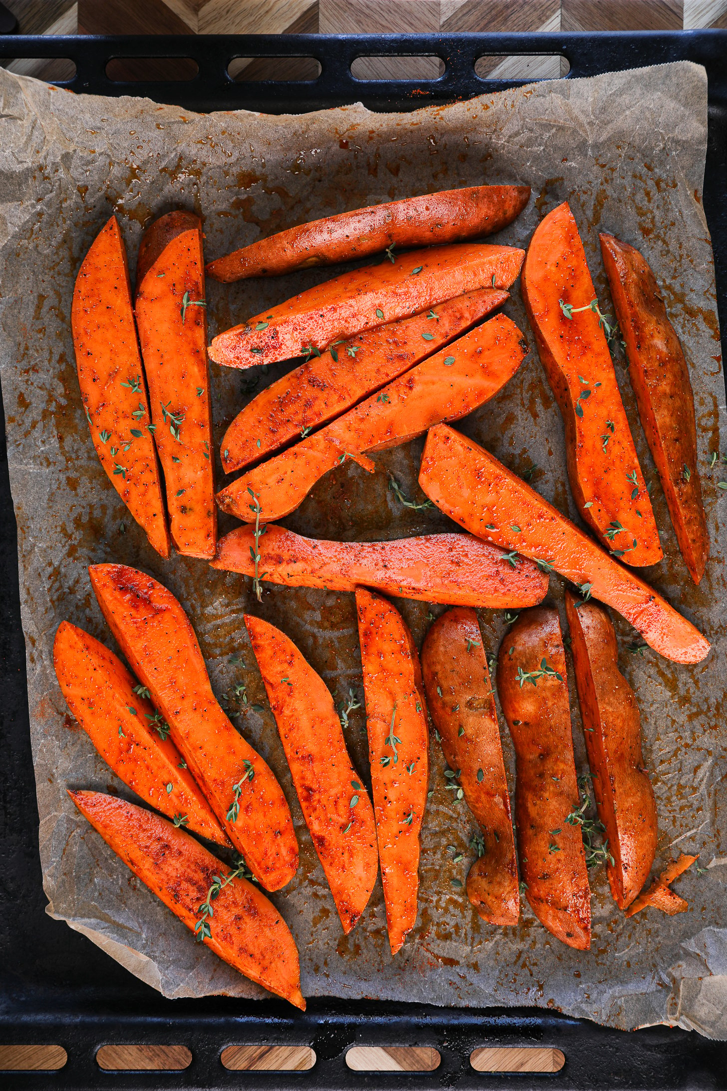 Raw sweet potato wedges, seasoned and drizzled with oil, adorned with fresh thyme leaves, resting on a lined baking sheet.