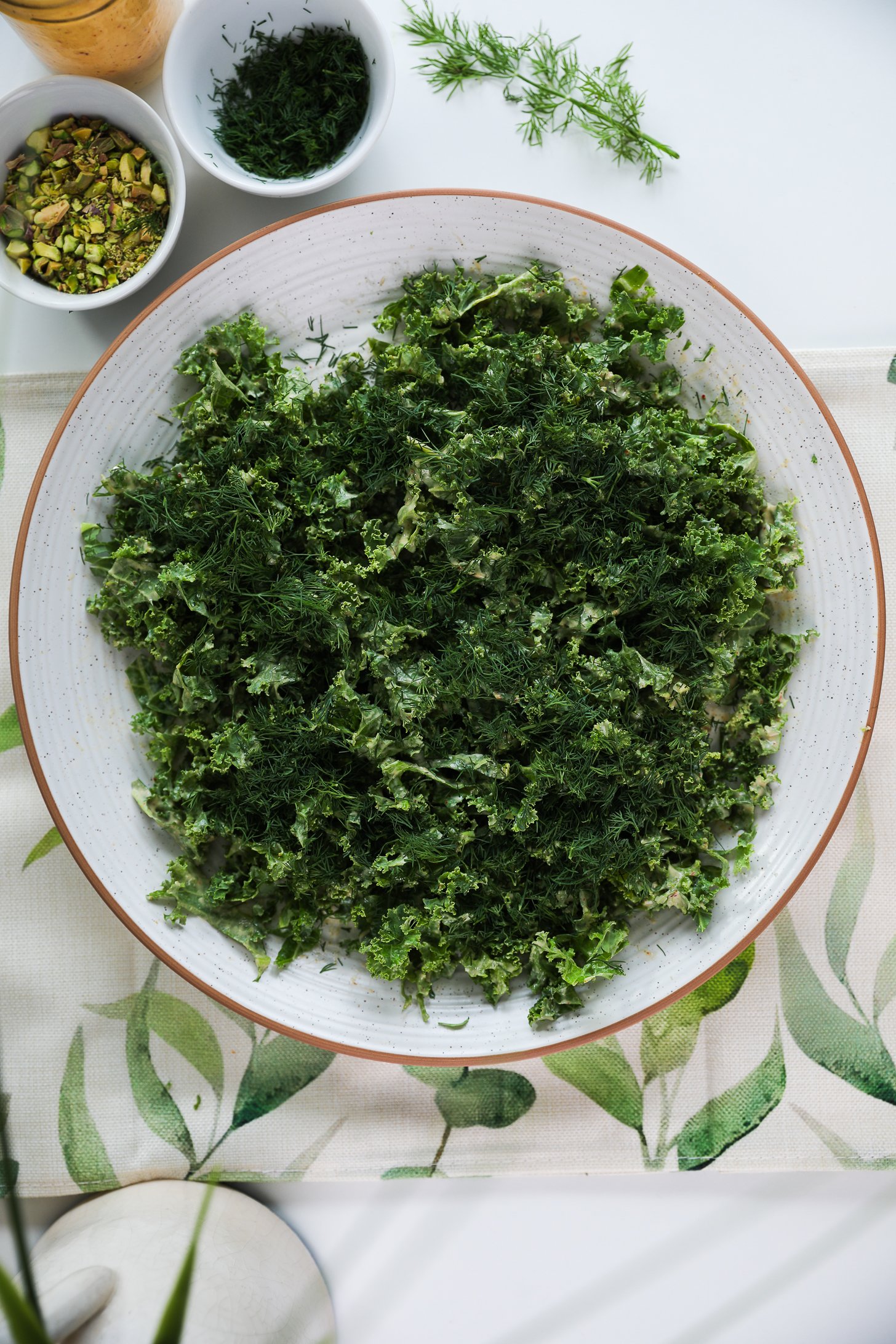 A large plate of chopped kale topped with fresh dill on a runner with ramekins of nuts and herbs nearby.