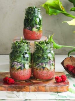 A vibrant styled display of three layered mason jar spring salads with raspberries and flowers in the fore- and backgrounds.