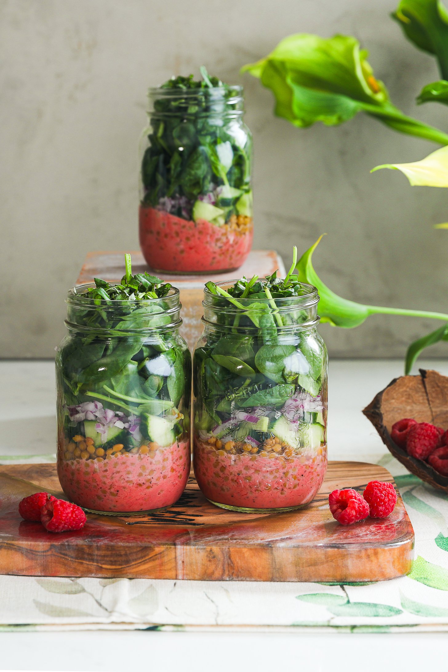 A vibrant styled display of three layered mason jar spring salads with raspberries and flowers in the fore- and backgrounds.