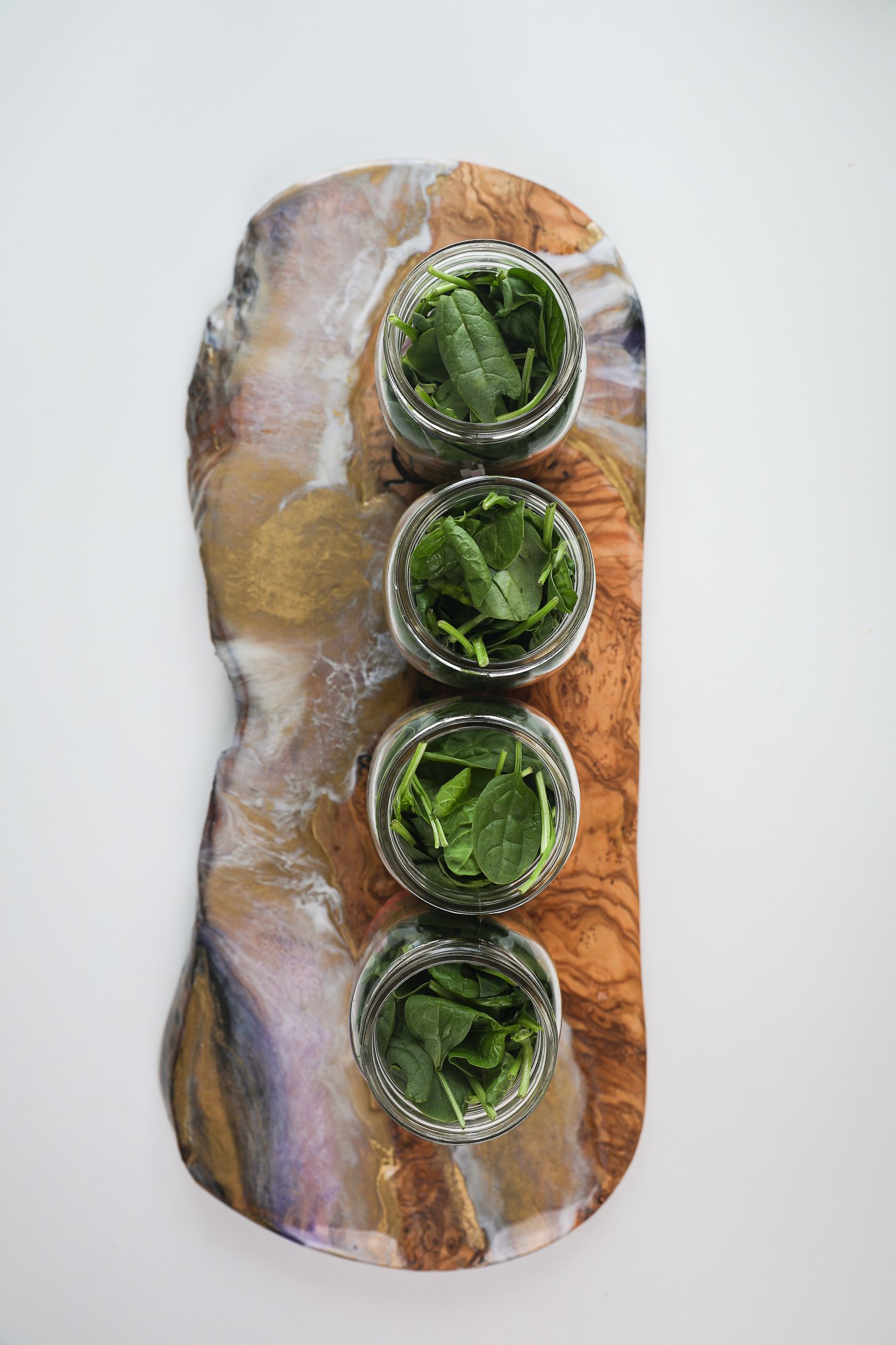Four mason jars lined up and filled to the rim with spinach placed on a long wooden board.