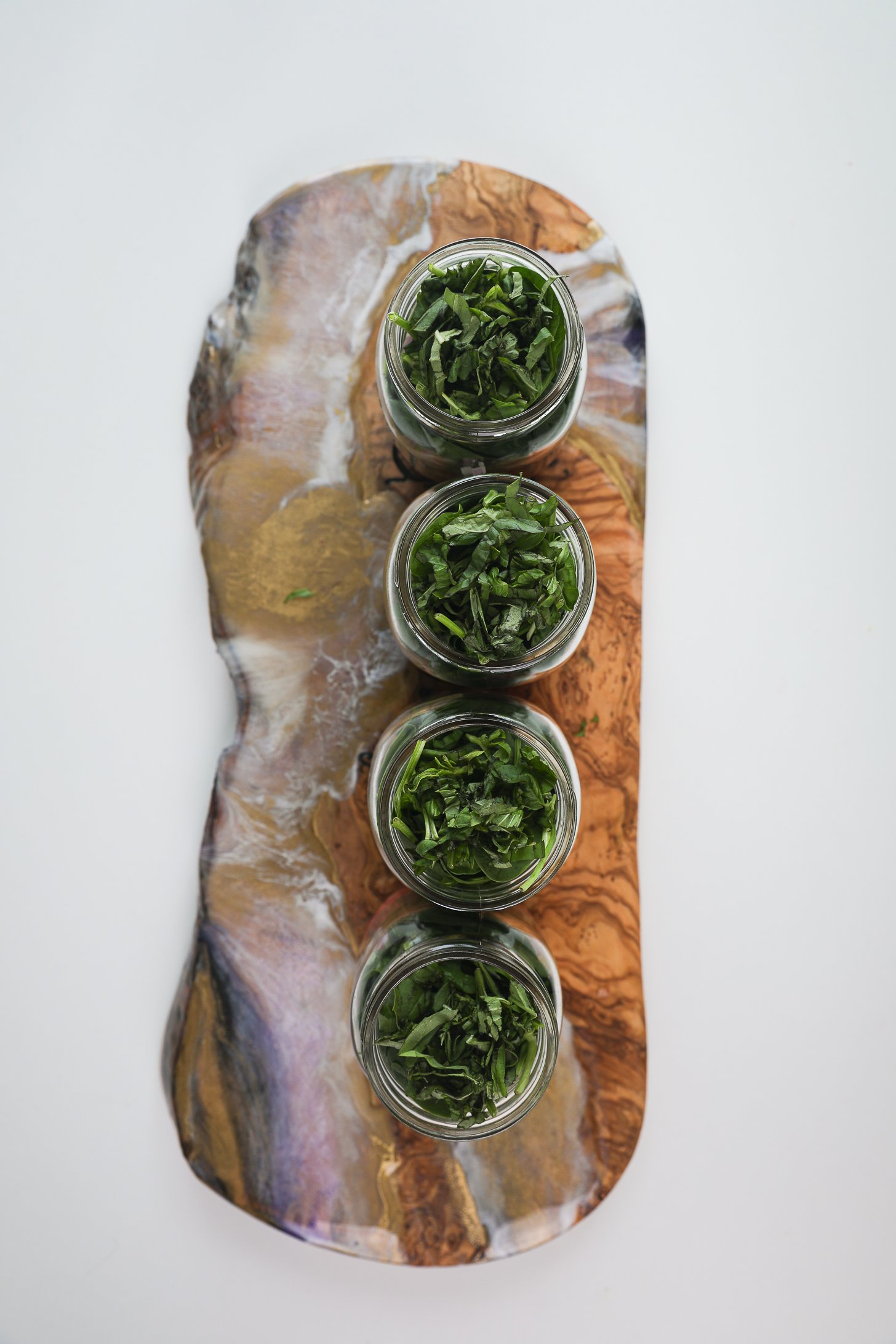 Four mason jars lined up and filled to the rim with chopped greens and basil placed on a long wooden board.