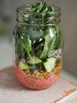 A face-on image of a mason jar containing a layered spring salad including a berry red dressing, spinach, cucumber and lentils.