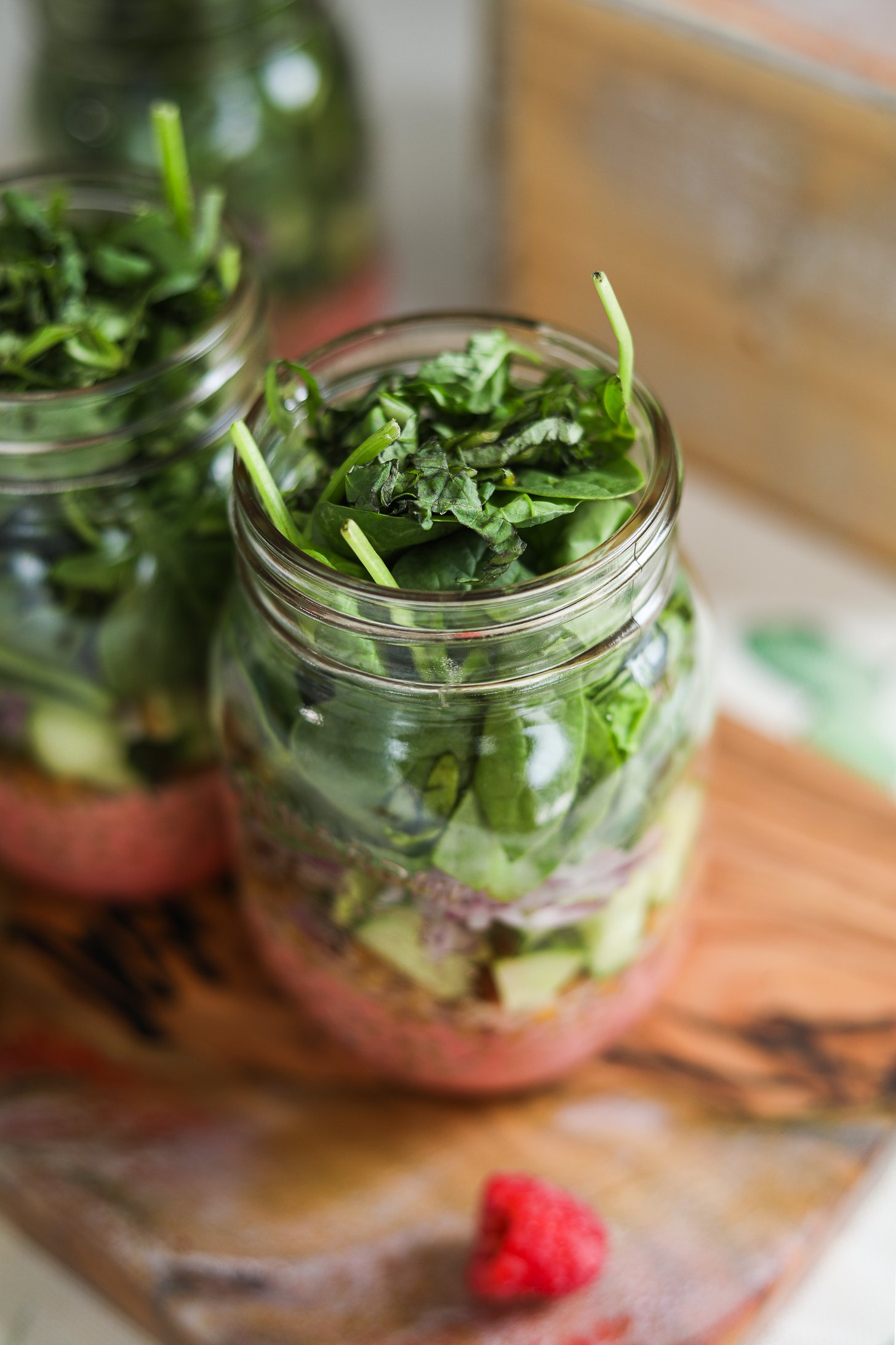 A perspective image of two mason jars containing a layered salad including a berry red dressing, spinach, cucumber and lentils.