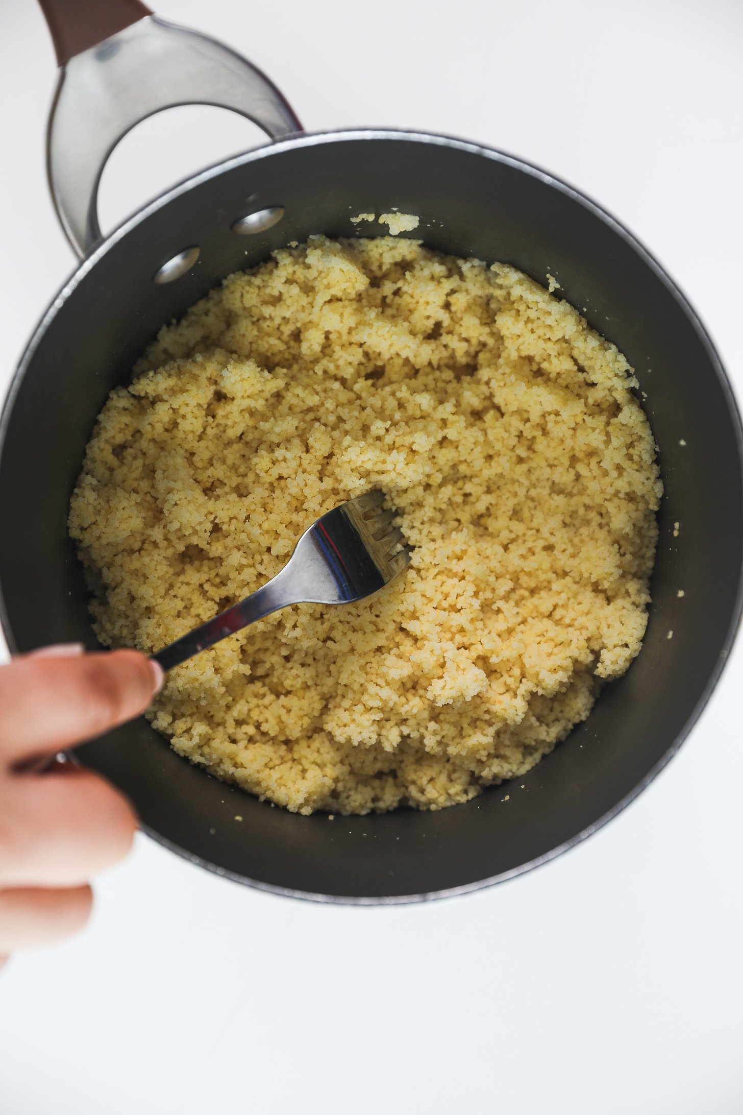 A hand holding a fork fluffing cooked couscous in a saucepan.