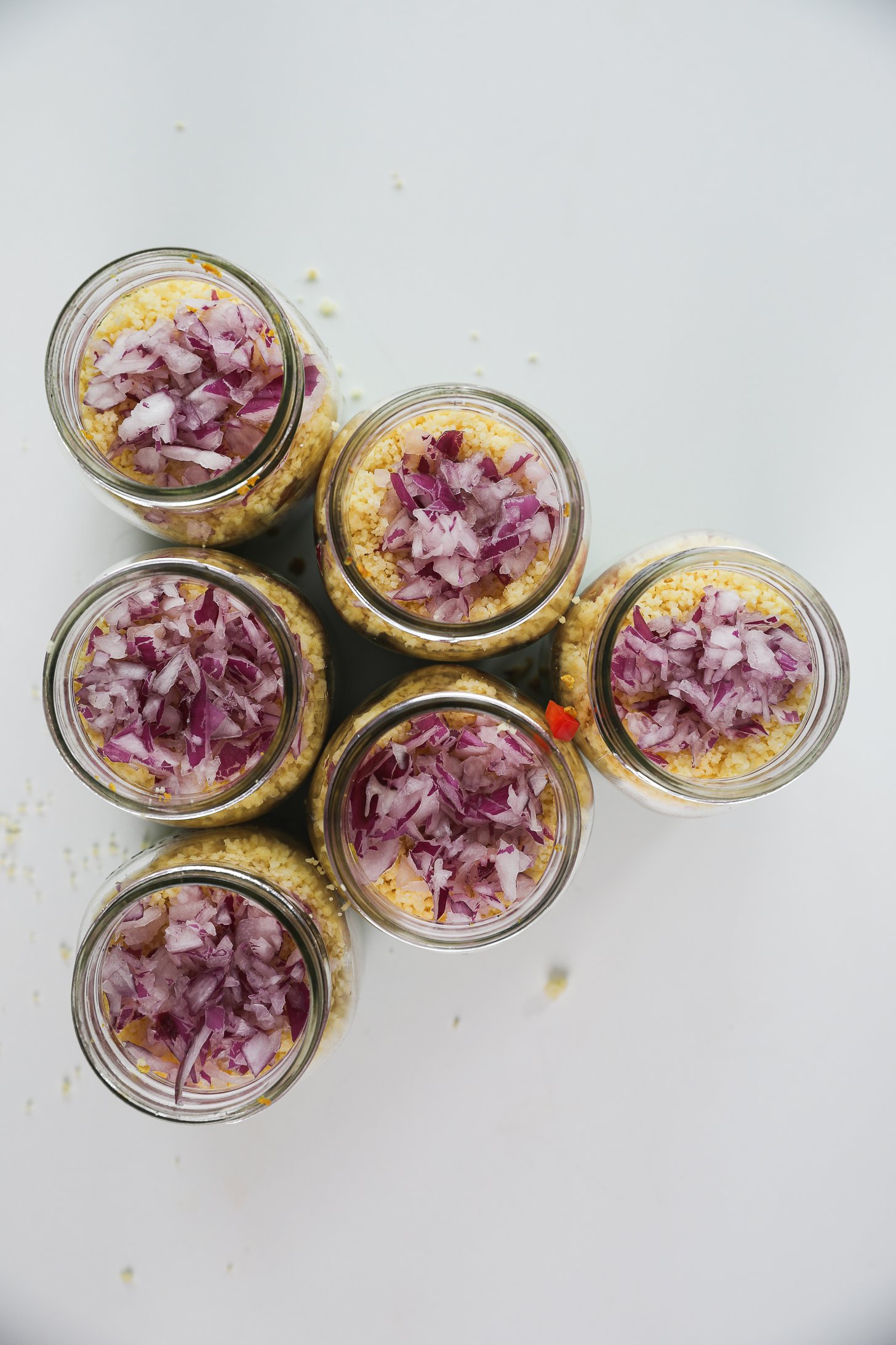 Six mason jars with avocado cilantro lime dressing at the base topped with a second layer of tandoori cauliflower, a third layer of chopped peppers, a fourth layer of couscous, and a fifth layer of chopped red onion.