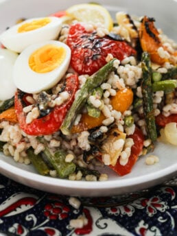 Perspective image of a bowl of barley salad with mixed grilled veggie topped with boiled egg halves.