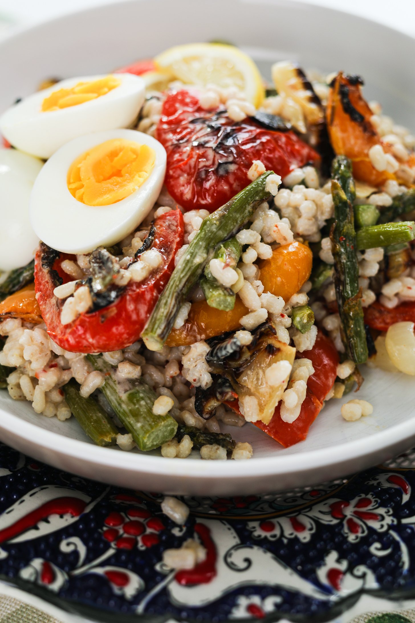 Perspective image of a bowl of barley salad with mixed grilled veggie topped with boiled egg halves.