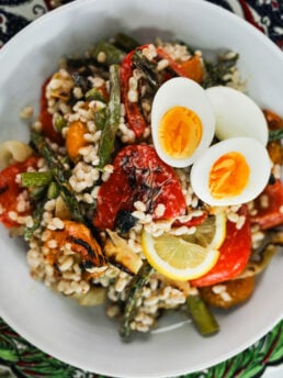 Top-view image of a bowl of mixed grilled vegetables, pearl barley in a creamy dressing topped with boiled egg halves.