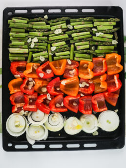 Top view of oven tray with chopped vegetables topped with garlic slices and oil.