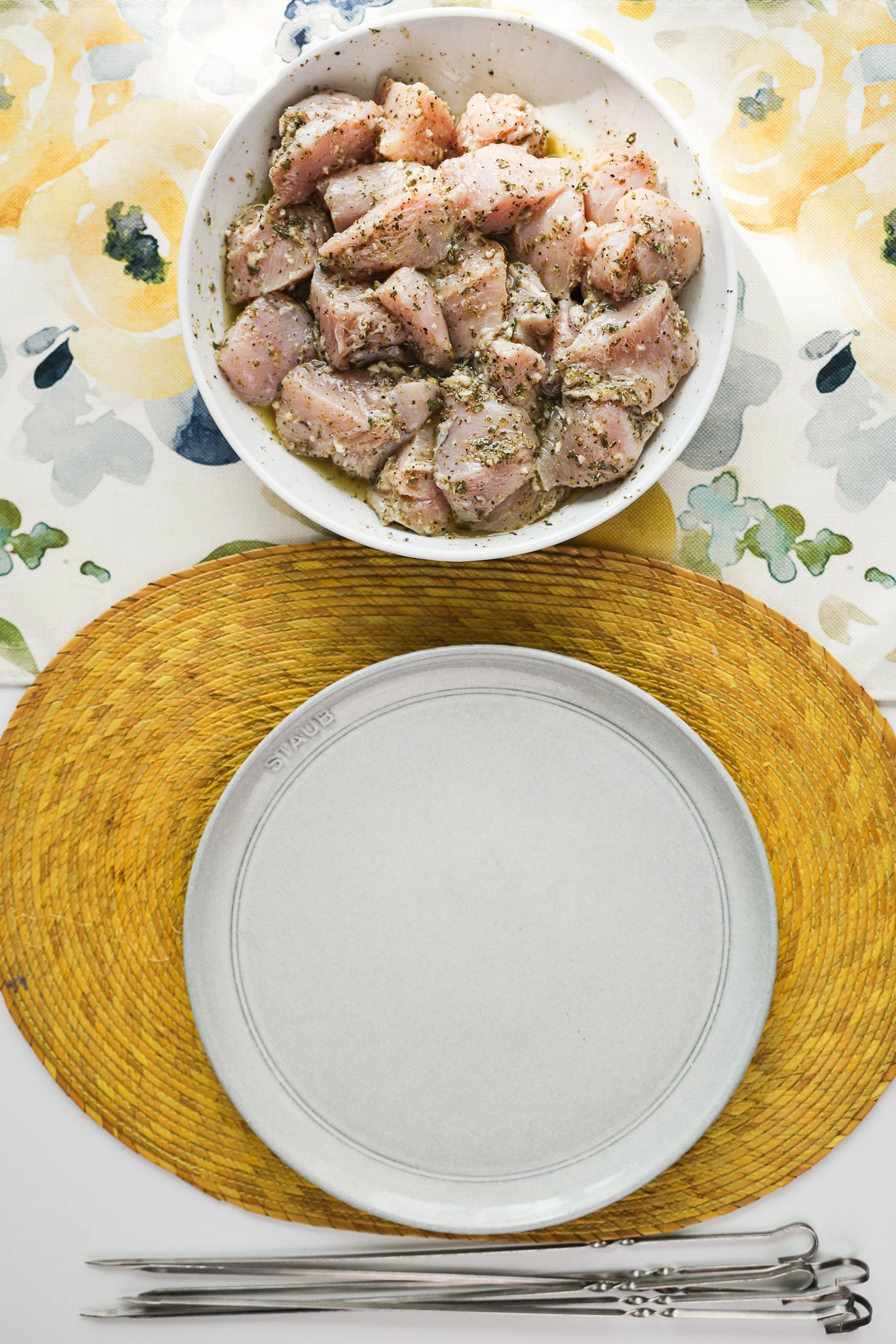 A bowl of marinated raw chicken breast next to an empty plate and metal skewers.