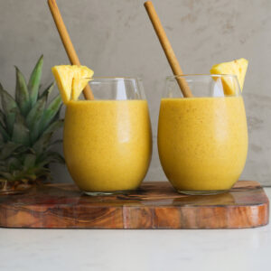 Perspective image of two glasses of yellow smoothie with bamboo straws in them.