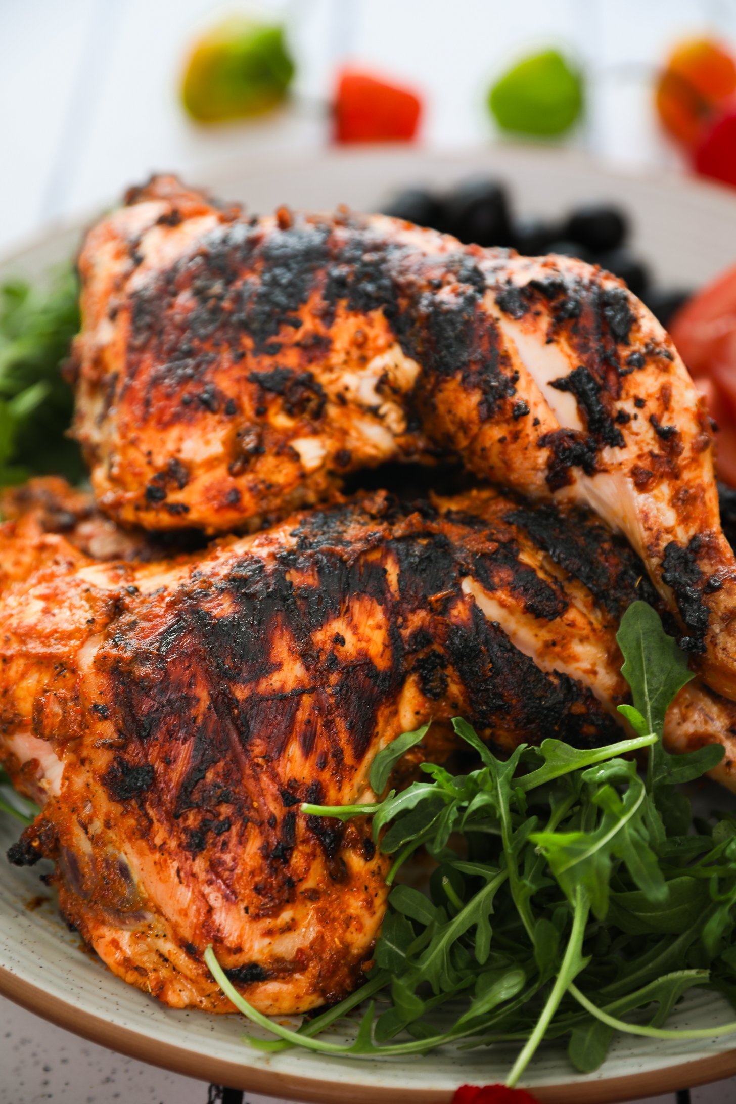 Perspective image of grilled chicken leg and thigh on top of arugula with colourful chillies in the background.