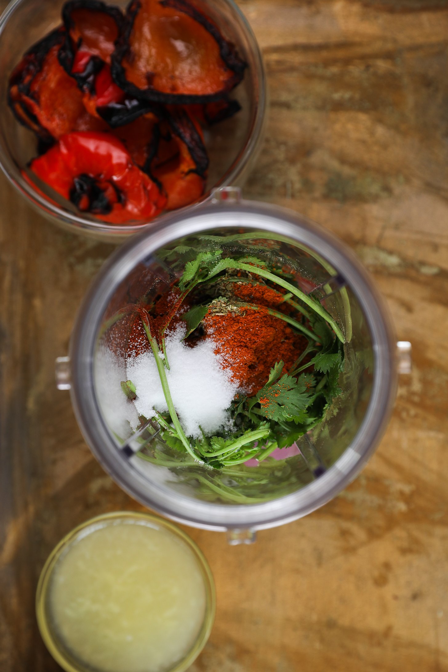 Top view of blender cup with cilantro leaves and stems and a red spice with salt with bowls of grilled peppers and lemon juice nearby.