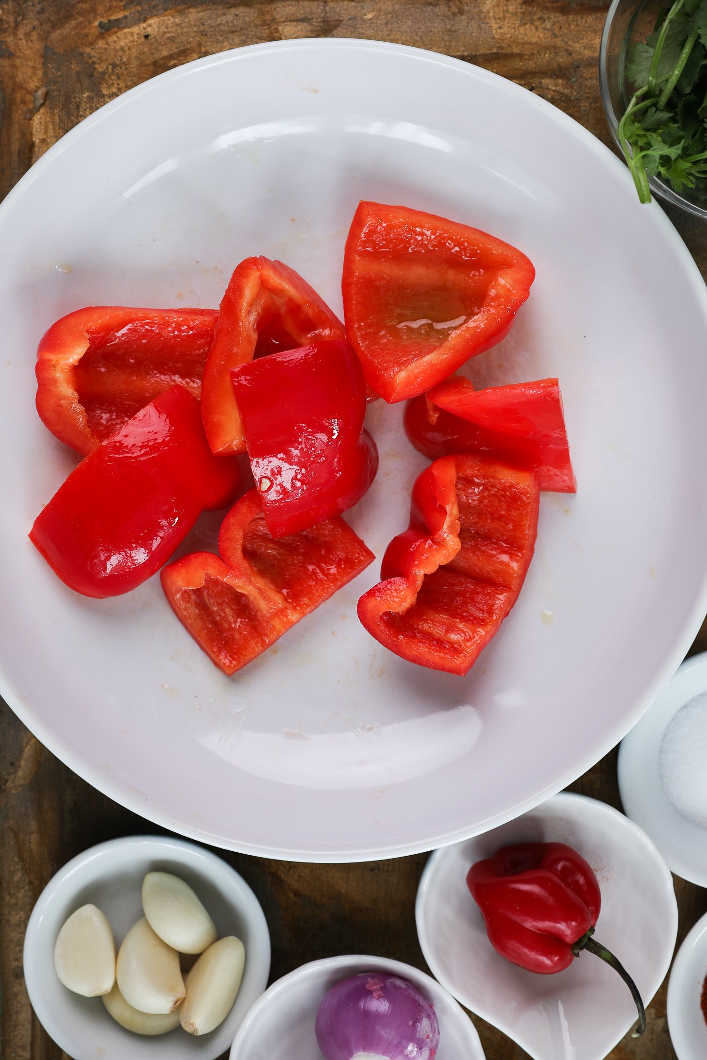 A white bowl of oil-coated red pepper chunks with small ramekins of garlic, shallot and chilli pepper nearby.