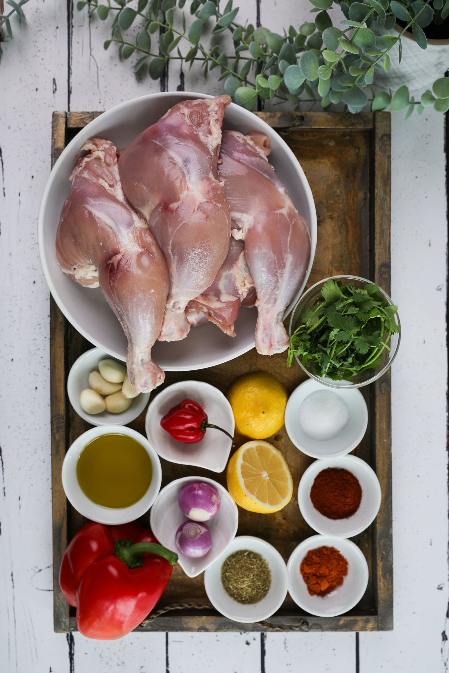 A selection of food ingredients including skinned chicken leg and thighs, spices, oil, red bell pepper, cilantro and a lemon half.