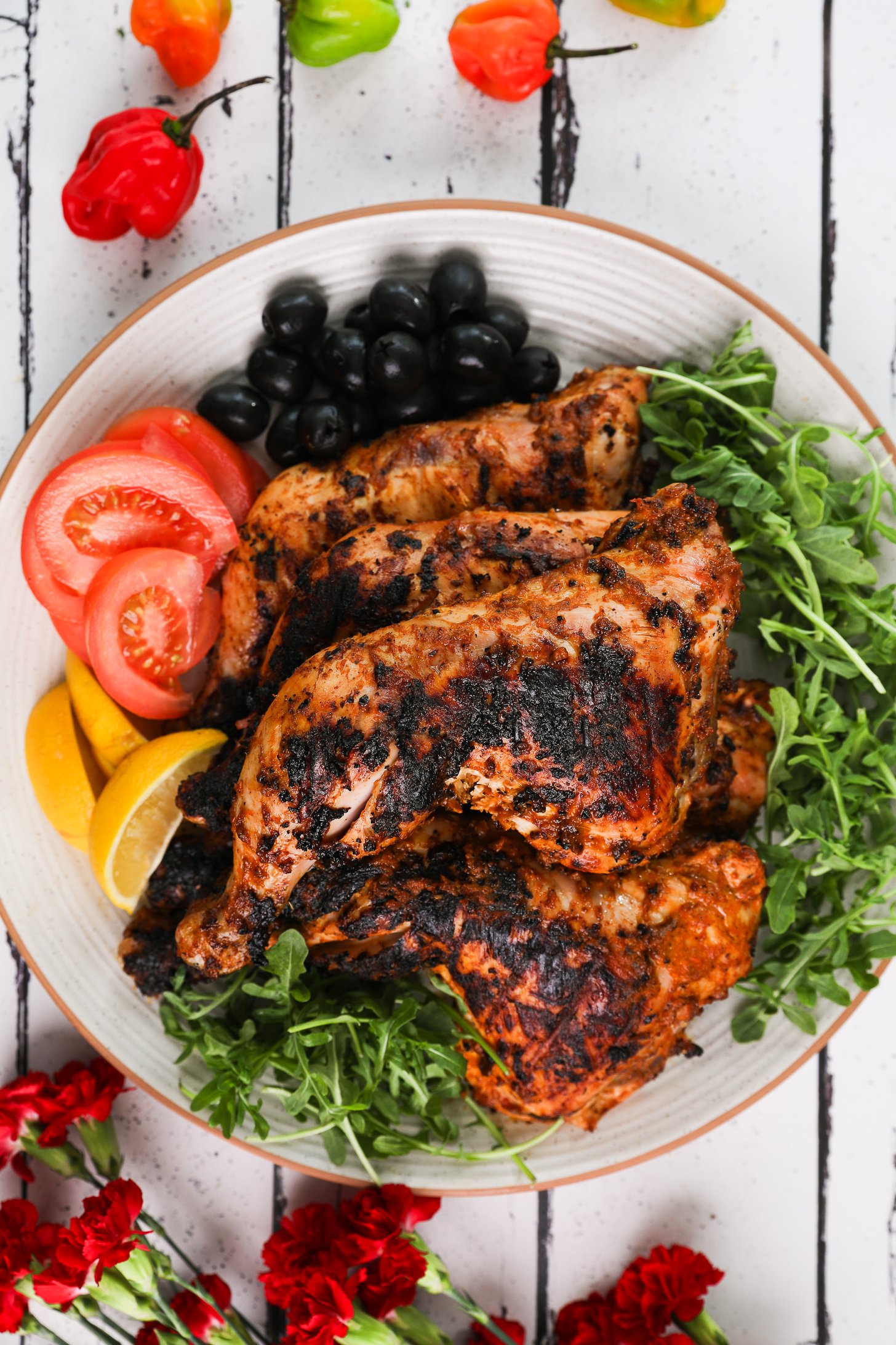 Top-down image of grilled chicken legs and thighs on a plate with arugula, tomato slices, lemon segments and olives.