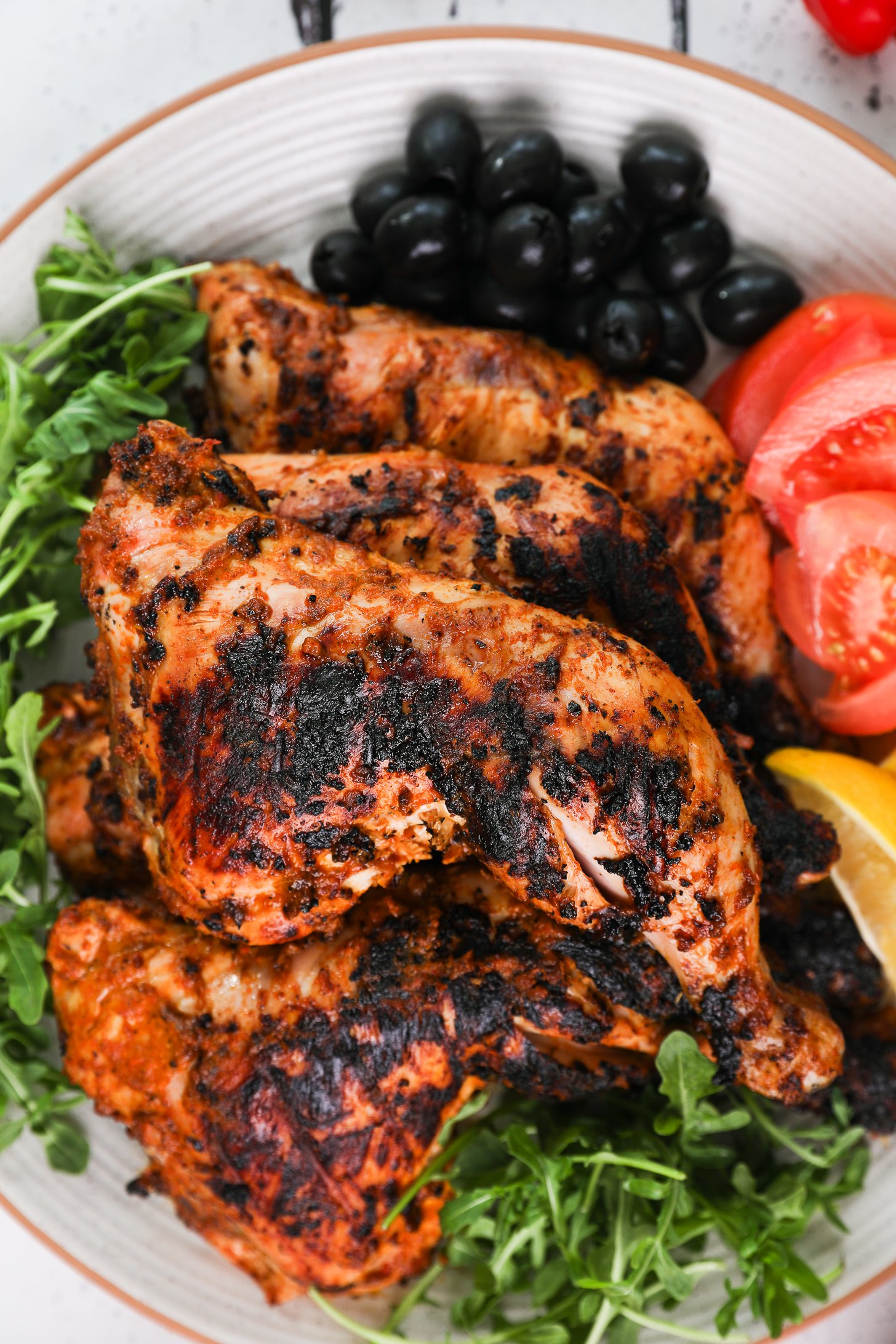 Top-down close up image of grilled chicken legs and thighs on a plate with arugula, tomato slices and olives.