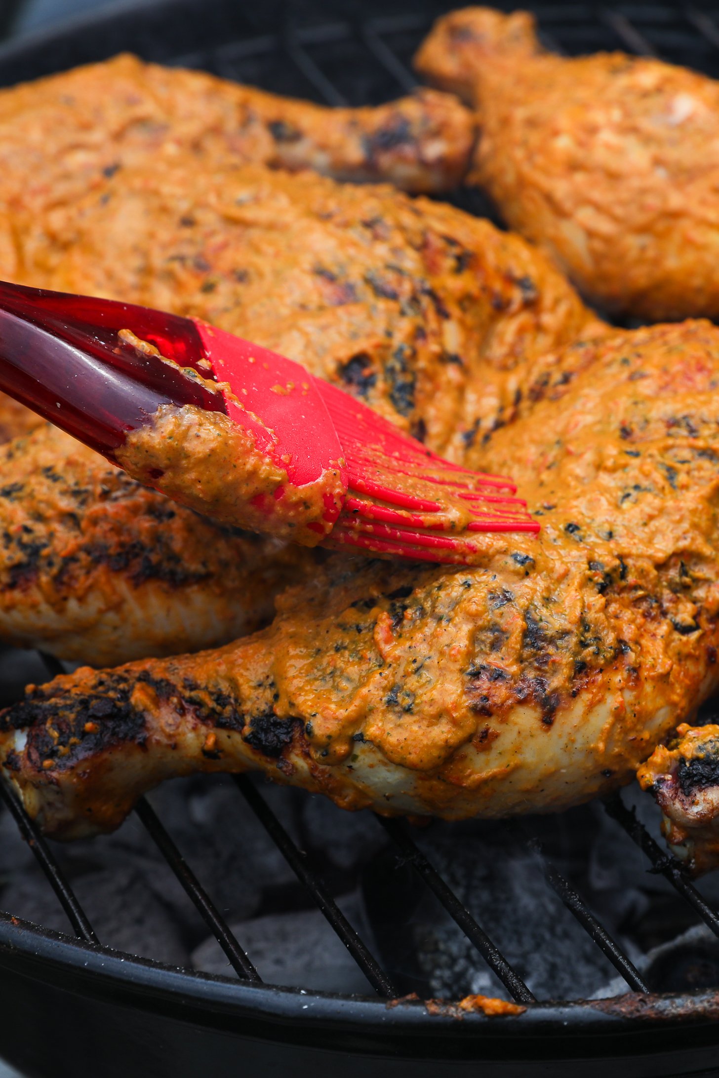 Brush basting marinade on the top side of a chicken leg and thigh on a charcoal grill with other marinated chicken legs and thighs.