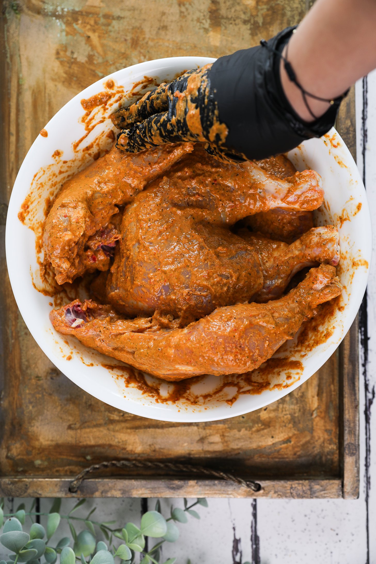 Hand wearing a black glove coating a pile of chicken legs and thighs with a thick orange marinade in a bowl.