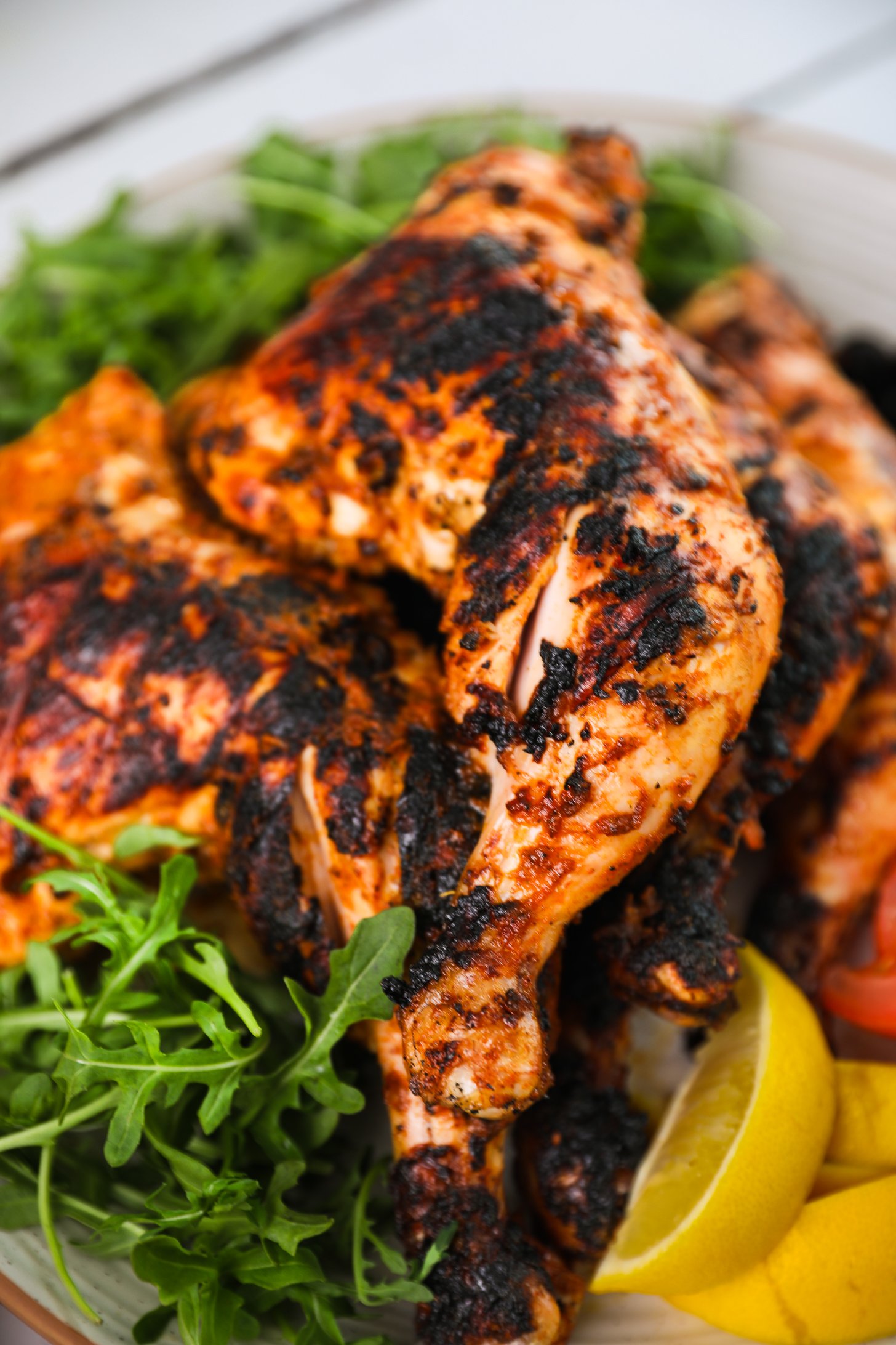 Perspective close up image of a pile of char-grilled spice coated chicken leg thighs atop arugula and lemon slices.