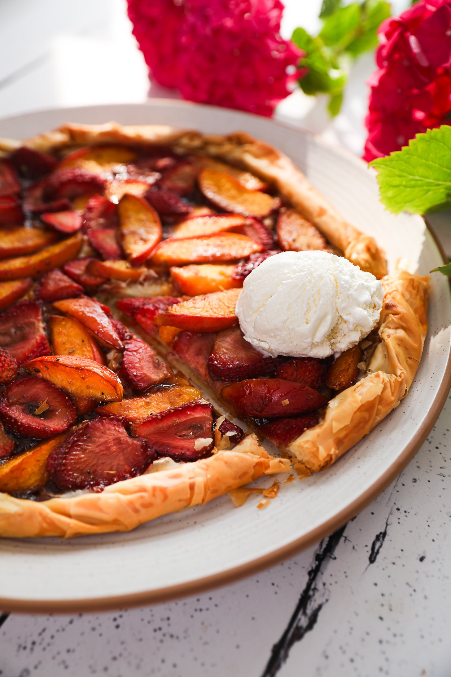 Perspective close up image of a strawberry and peach galette with a filo pastry base, topped with a scoop of ice cream on a cut slice. There are pink flowers in the background.