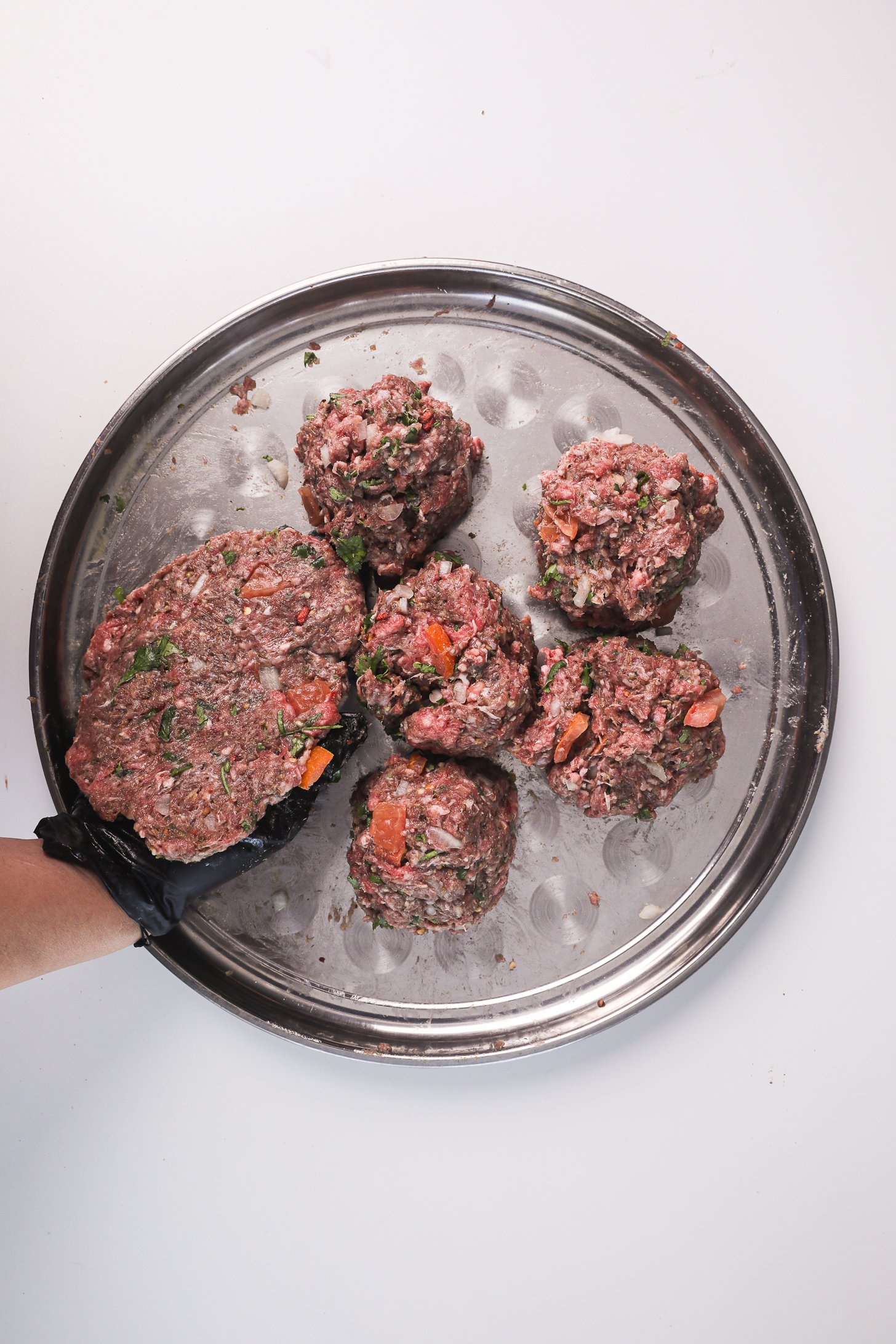 A gloved hand flattening a Chapli Kebab ball into a patty, with other raw kebab balls nearby on the tray.