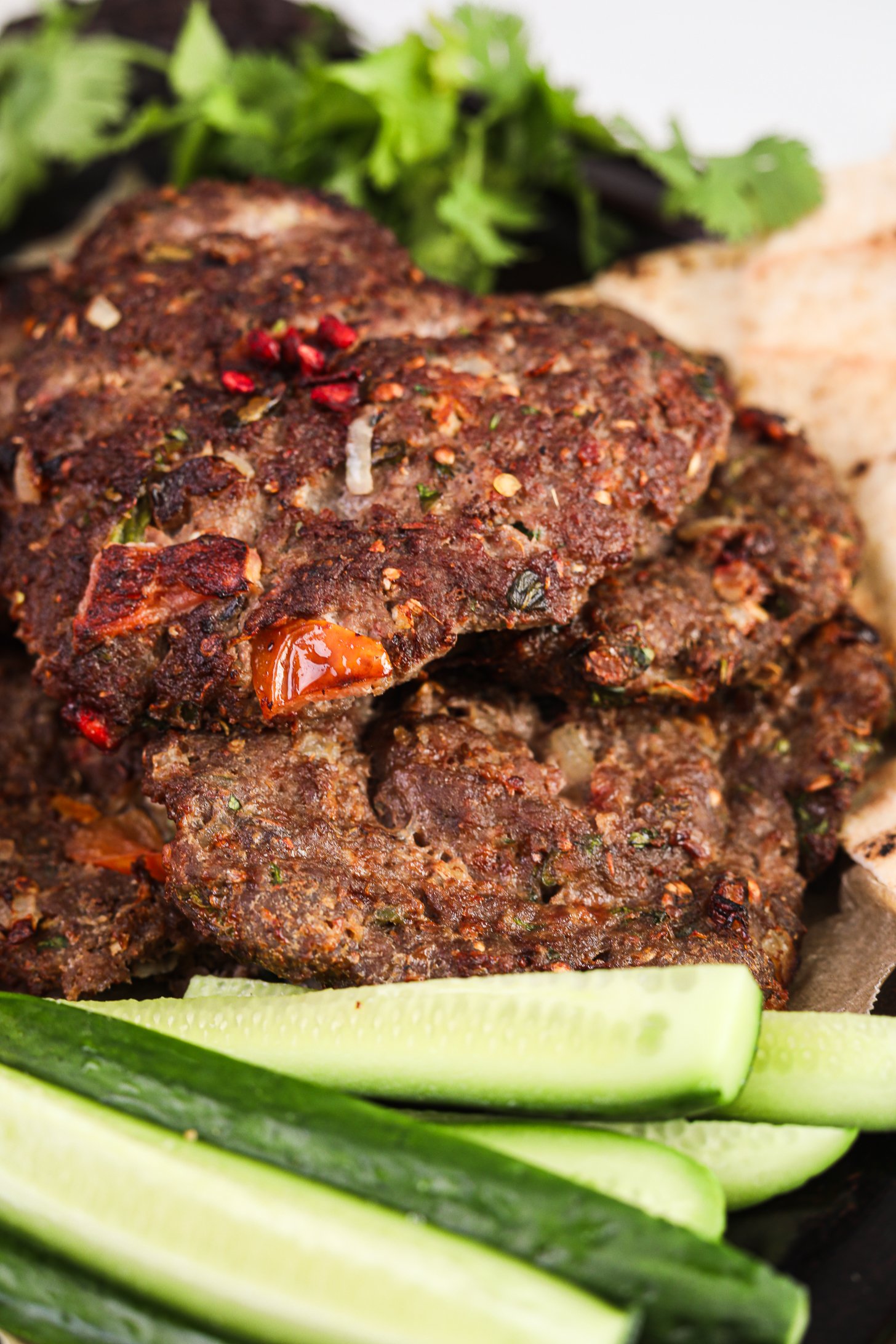 Close-up of Chapli Kebabs with visible chunks of tomatoes, served with cucumbers and coriander.