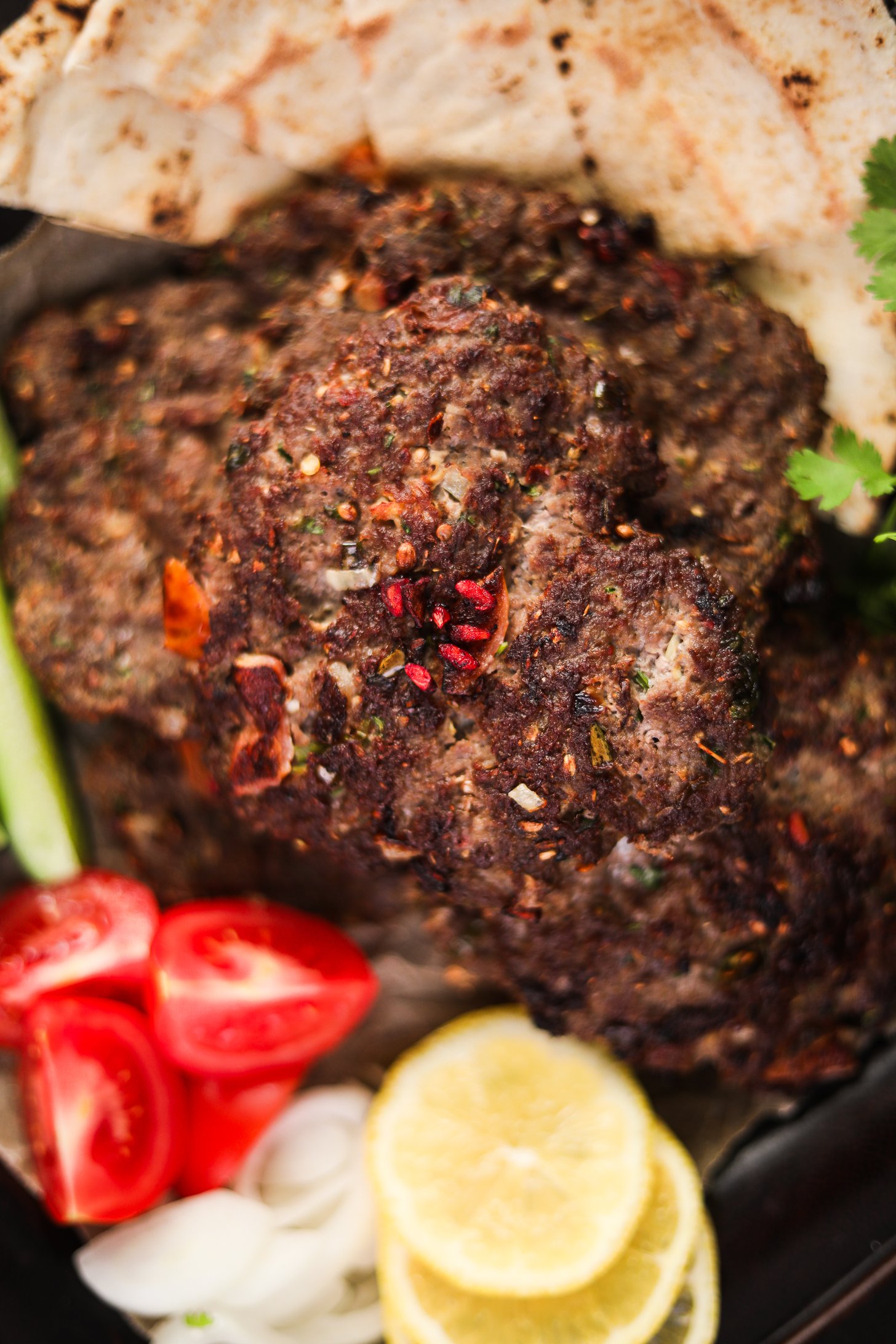 Top view of Chapli Kebabs on a tray with naan, cucumber, tomatoes, onions, and lemon slices.