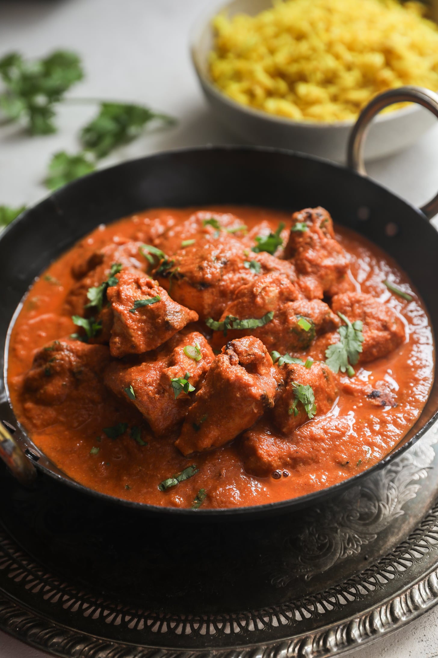 Un plateau de service noir rempli de tomates au curry crémeuses et de morceaux de poitrine de poulet, garni de coriandre hachée, accompagné d'un bol de riz jaune.